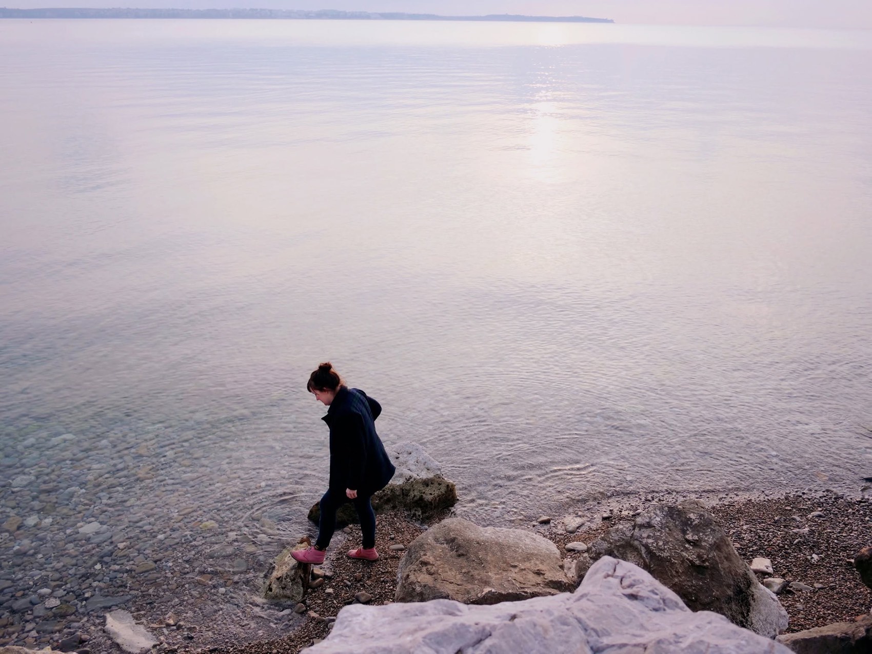 Sarah walking along a pebbled shoreline at dusk, still water stretching to the horizon