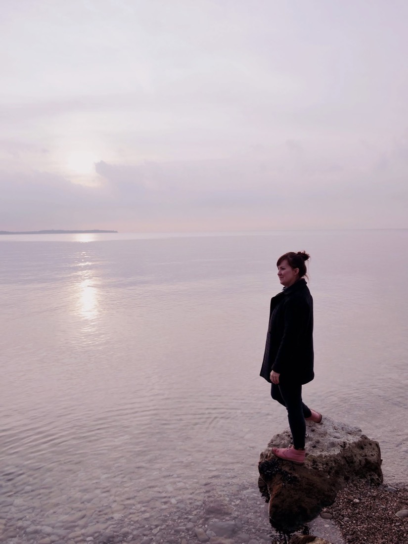 Sarah standing on a rock at the water's edge, looking out across a still sea at dawn